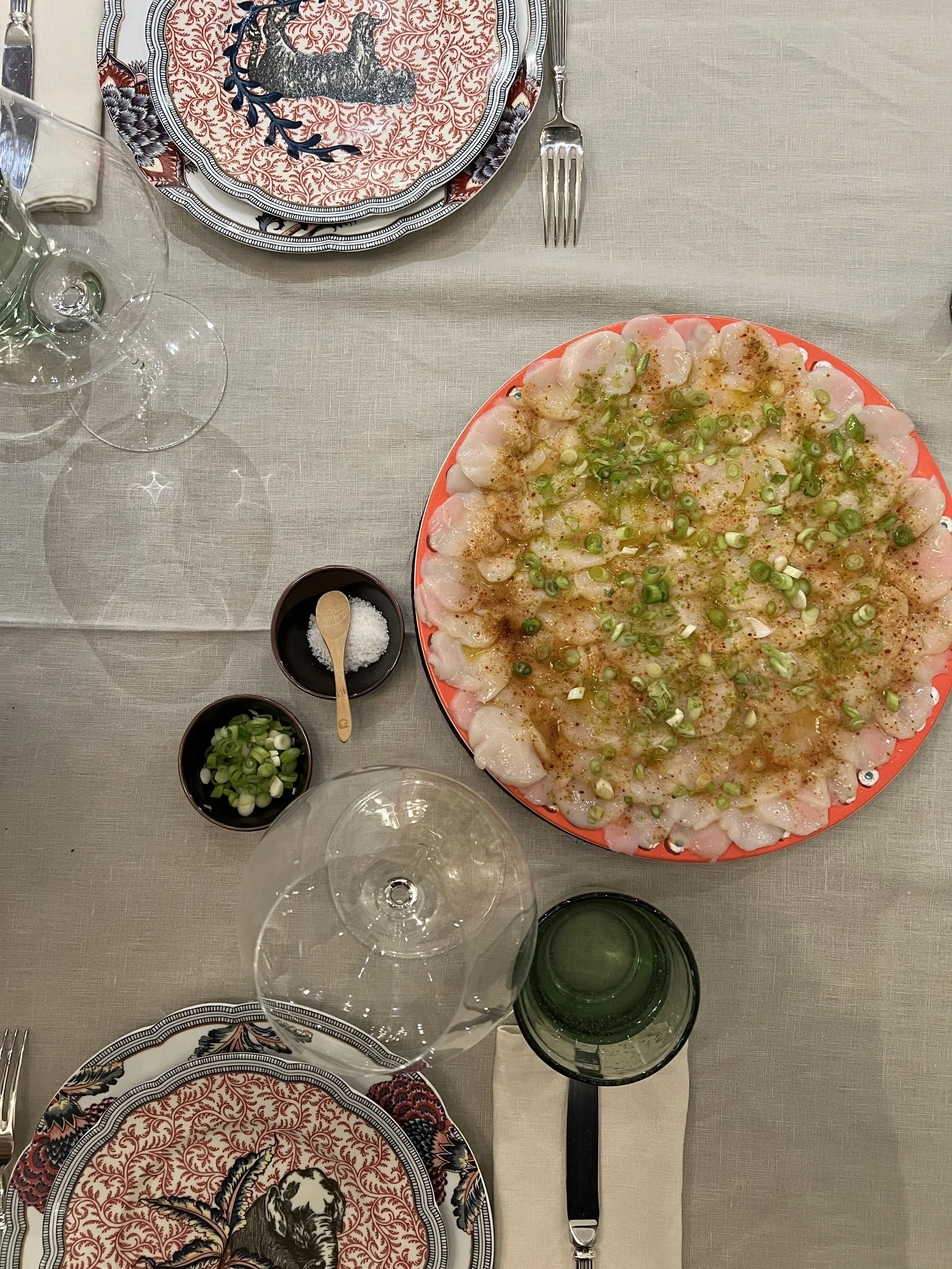 A table set with a large plate of sashimi garnished with green onions and seasonings, a smaller dish of salt, a bowl of chopped green onions, empty wine glasses, a green glass bottle, and decorative plates with a lion and an elk design.
