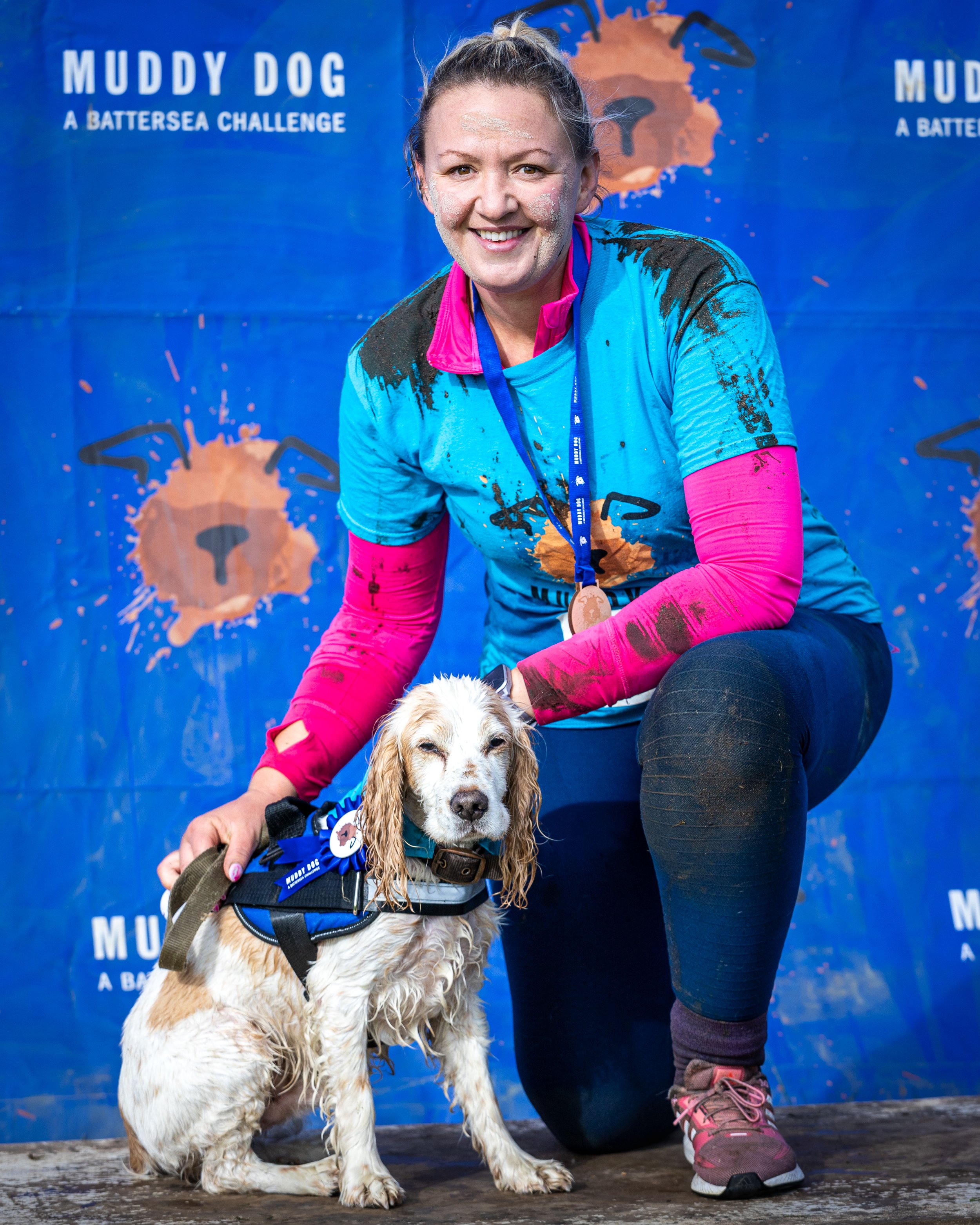 A woman in a blue and pink athletic outfit kneeling next to a dog with a medal, in front of a blue banner that says 'Muddy Dog A B.A.T.T.E.R.S.E.A. Challenge'.