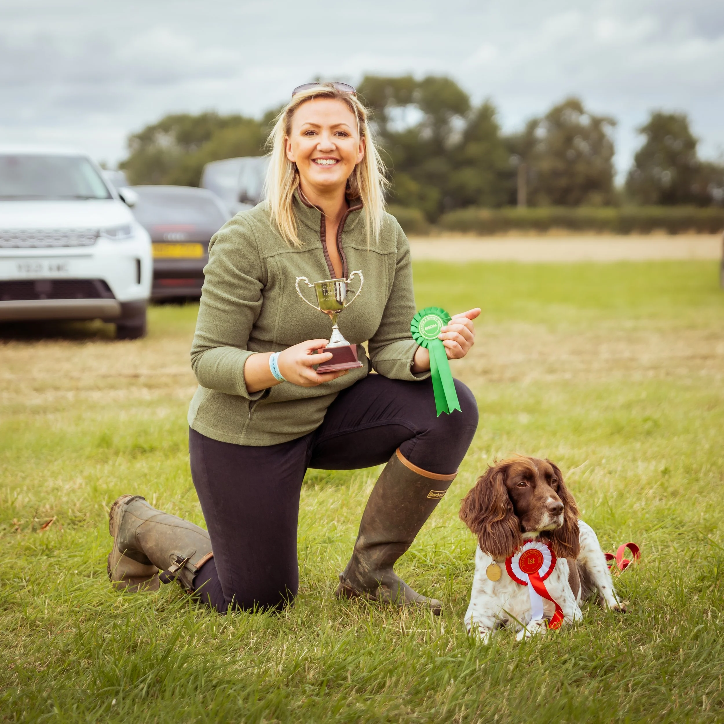 A woman kneeling on grass, holding a trophy and a green award ribbon, smiling. Beside her is a dog with a red ribbon and medal, sitting on the grass. In the background, there are cars and trees under a cloudy sky.