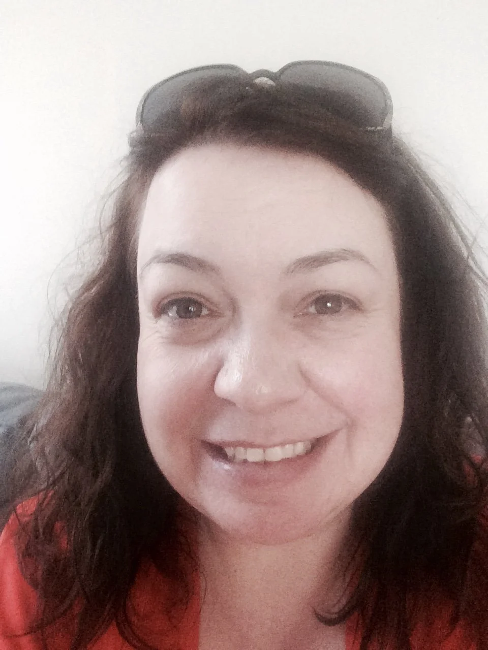 A smiling woman with dark wavy hair, wearing sunglasses on her head and a red top, in front of a plain white background.