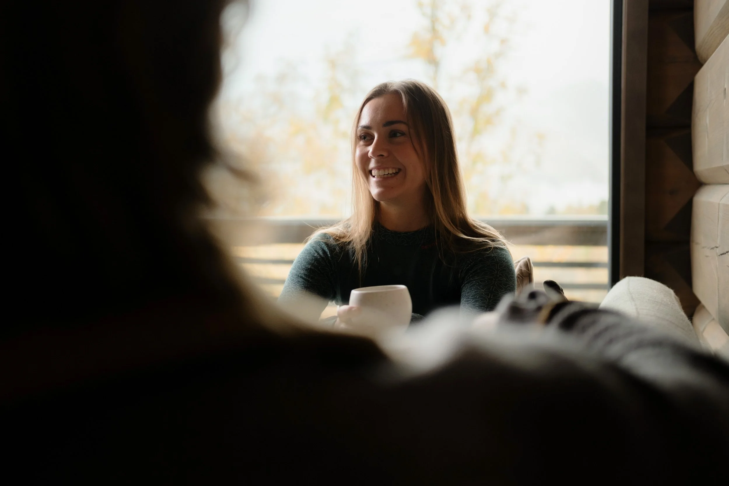 Smiling woman holding a mug indoors, with blurred foreground and a window in the background.