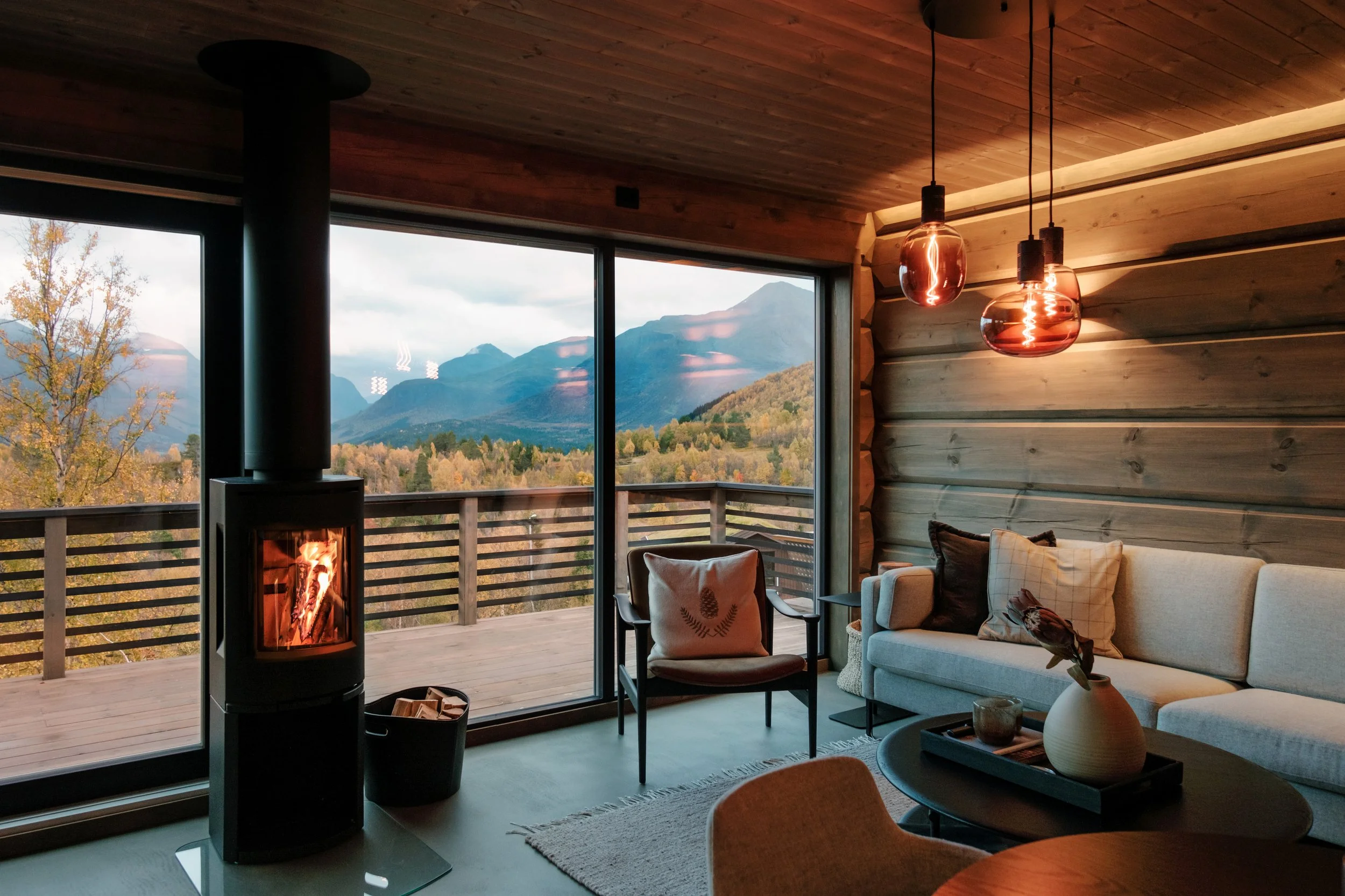 Cozy wooden living room interior with fireplace, large windows overlooking mountains, sofa, chair, table, and pendant lights.