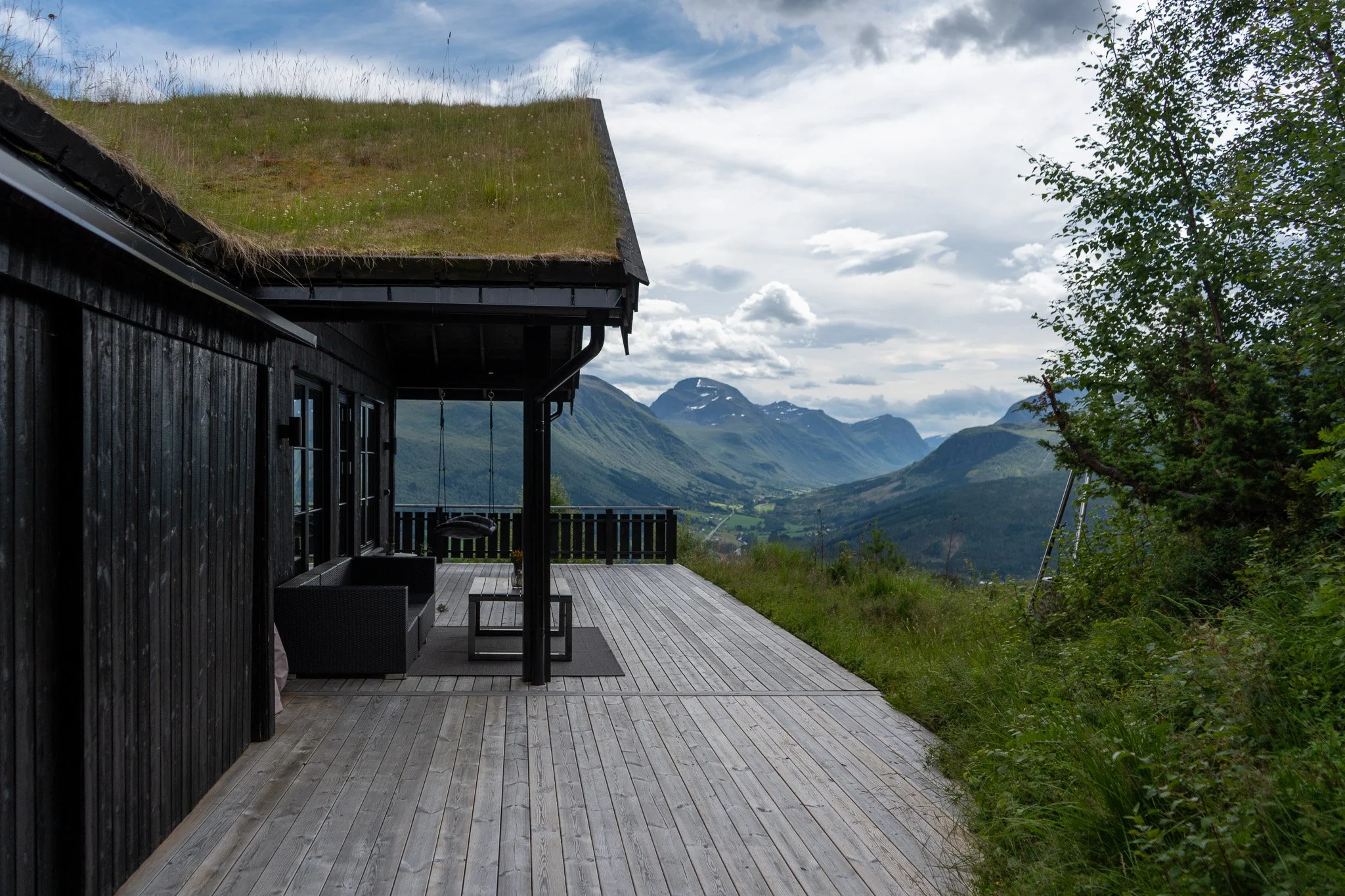 Wooden cabin with grass roof on a deck overlooking a mountain valley with trees and cloudy sky.