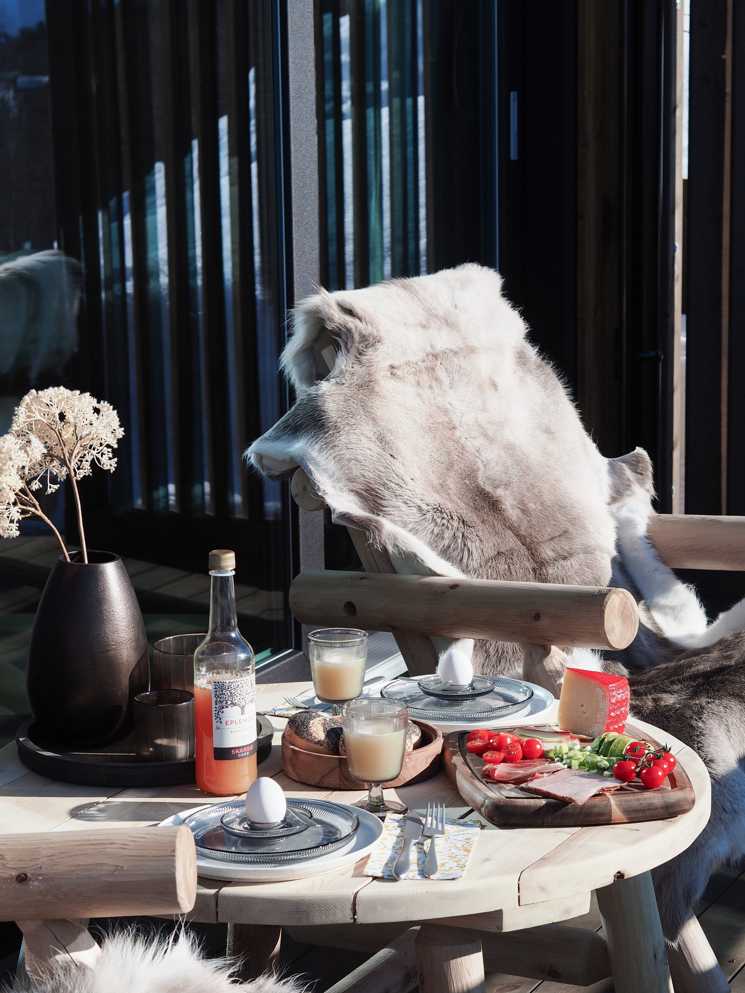 Rustic outdoor breakfast table setup with wooden chairs and fur drapes. The table features a bottle of juice, two glasses with a light beverage, a vase with dried flowers, a hard-boiled egg, and a platter with tomatoes, cucumber slices, cheese, and d