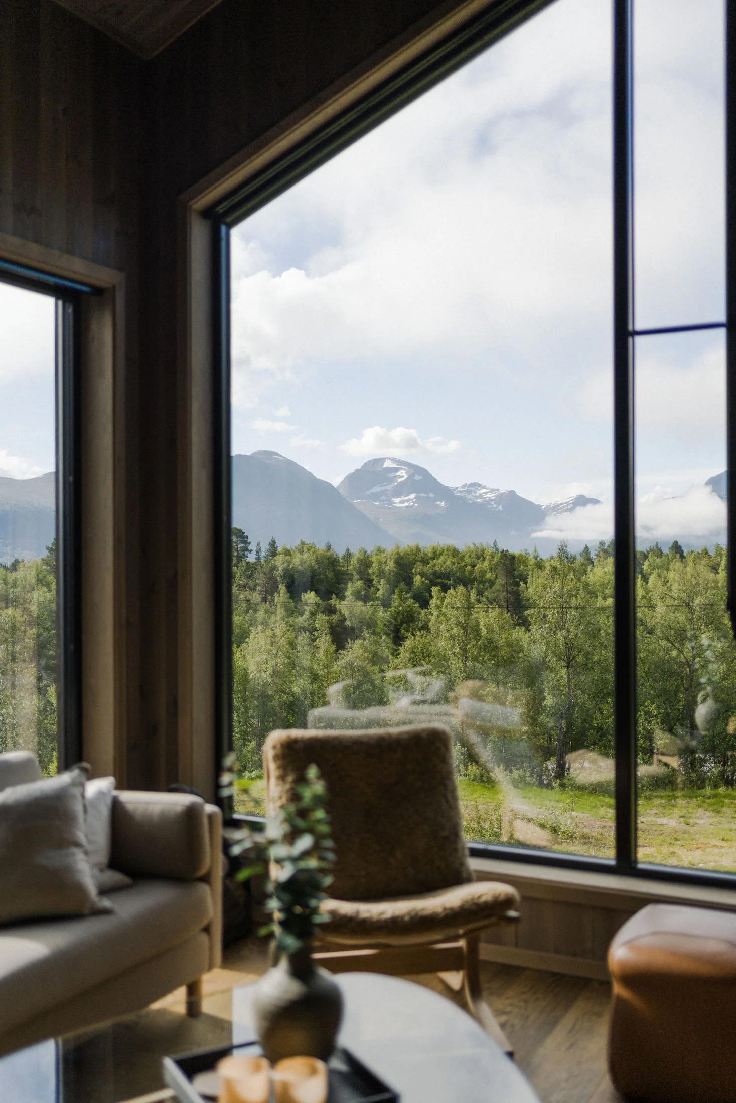 Interior of a modern living room with large windows showing a scenic view of mountains and forest. The room features a sofa, a wooden chair, and a coffee table with a plant and candles.