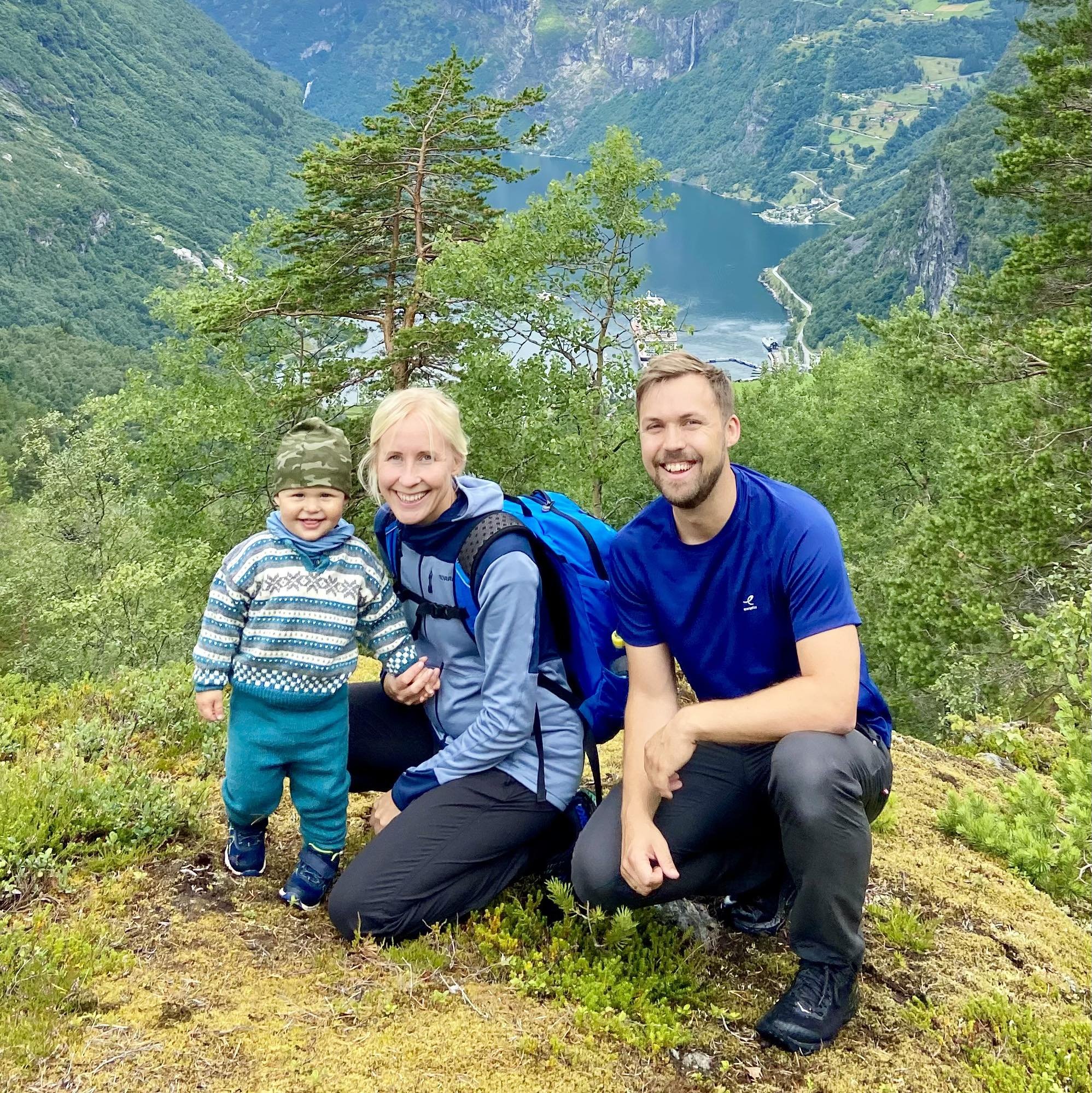 A family of three posing on a scenic mountain trail with lush greenery and a fjord in the background. A small child in a patterned sweater is smiling between two adults. The woman wears a blue jacket with a backpack, and the man is in a blue shirt. They are surrounded by trees and plants.