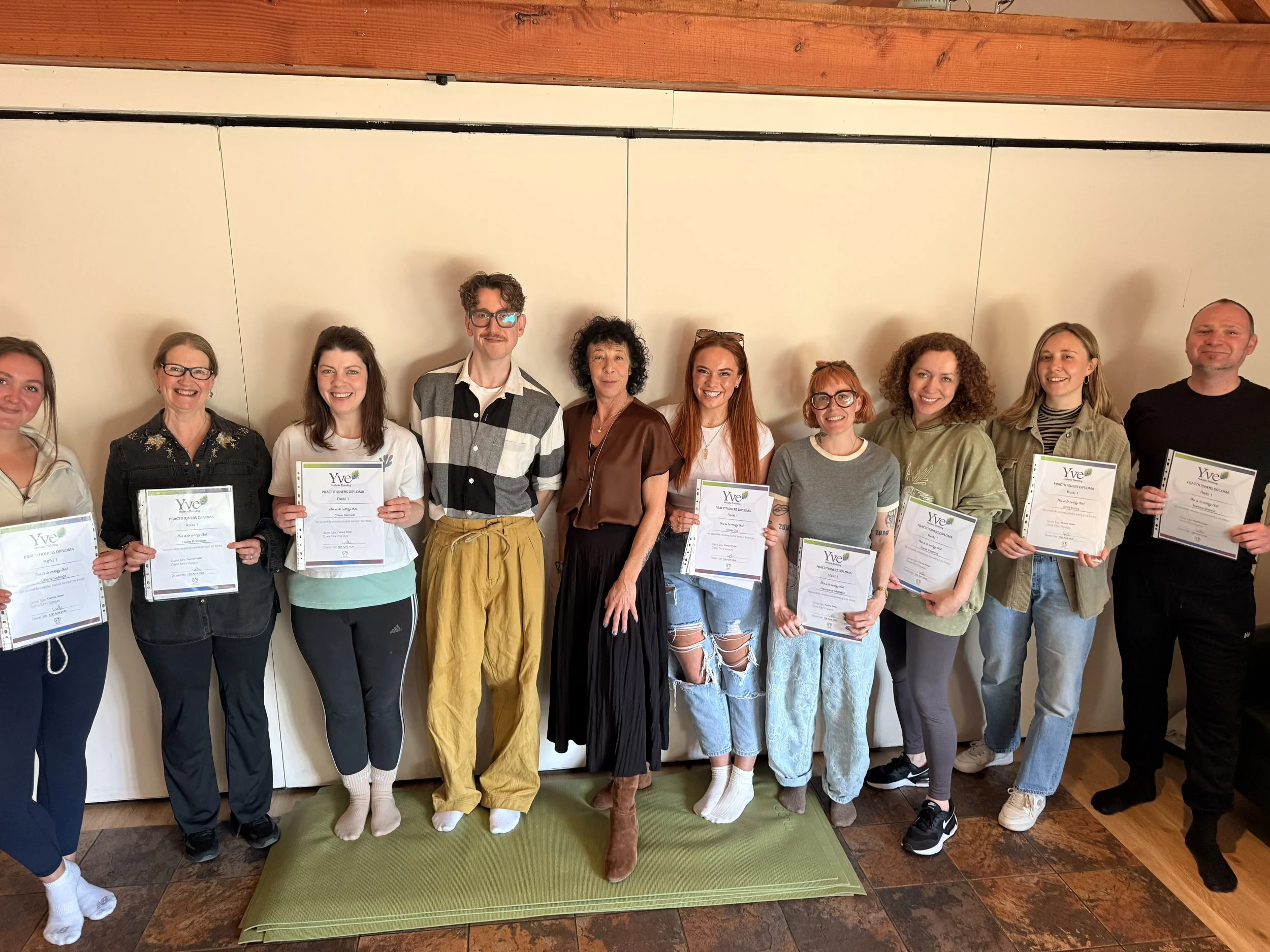 Group of ten people standing in a row against a plain wall, some holding certificates, dressed casually, smiling, in an indoor setting.
