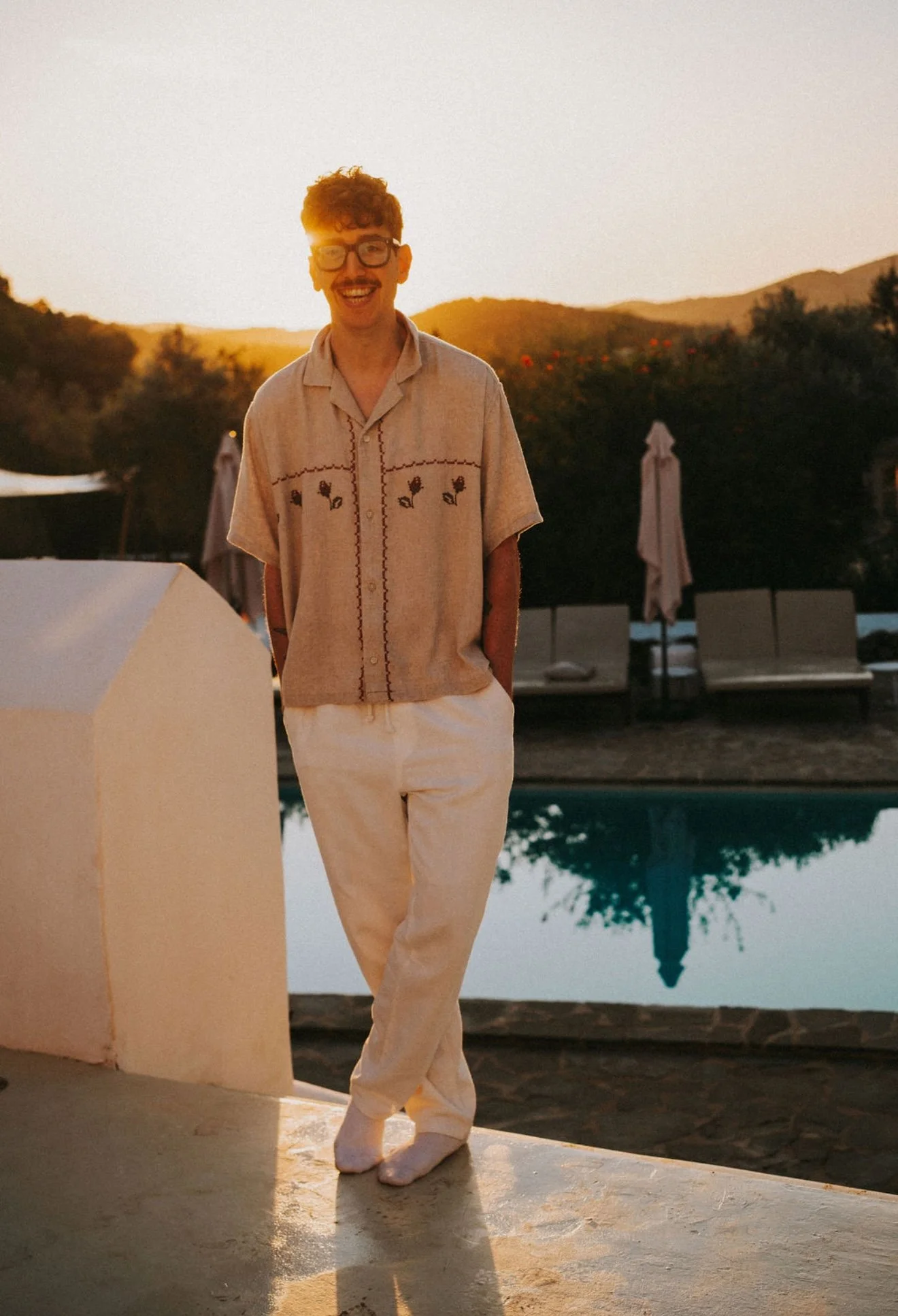 A man with curly hair, glasses, and a mustache smiling at sunset outdoors. He's wearing a beige embroidered shirt and cream-colored pants, standing near a pool with lounge chairs and umbrellas in the background.