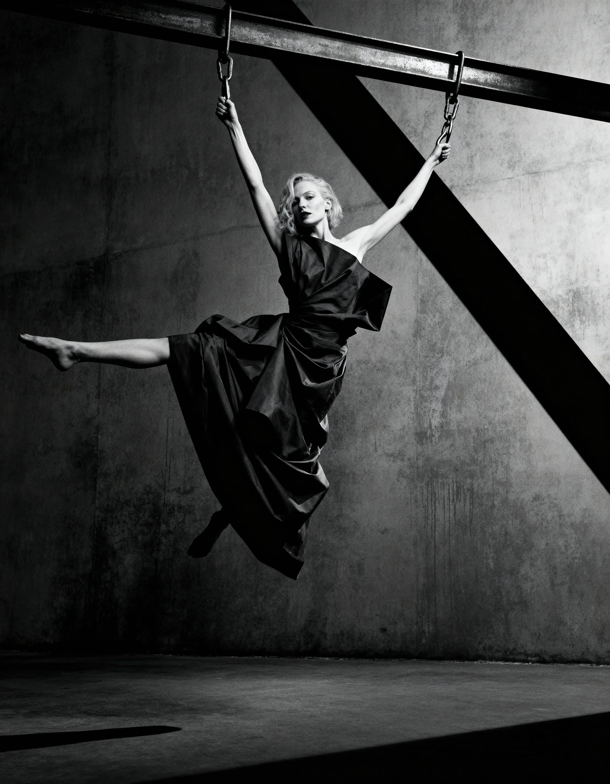 A black-and-white photograph of a woman in a dark dress hanging from chains in an industrial setting. She is suspended in mid-air with one arm holding a chain and one leg extended outward. The background consists of a concrete wall and floor with stark shadows.