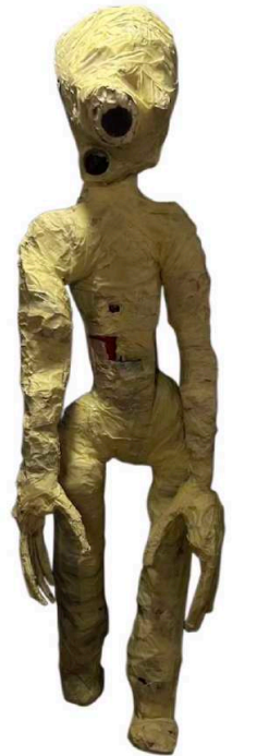 Statua di un essere umanoide con testa grande e occhi neri, fatta di carta o cartapesta, con un aspetto disorganizzato e in stile artistico.
