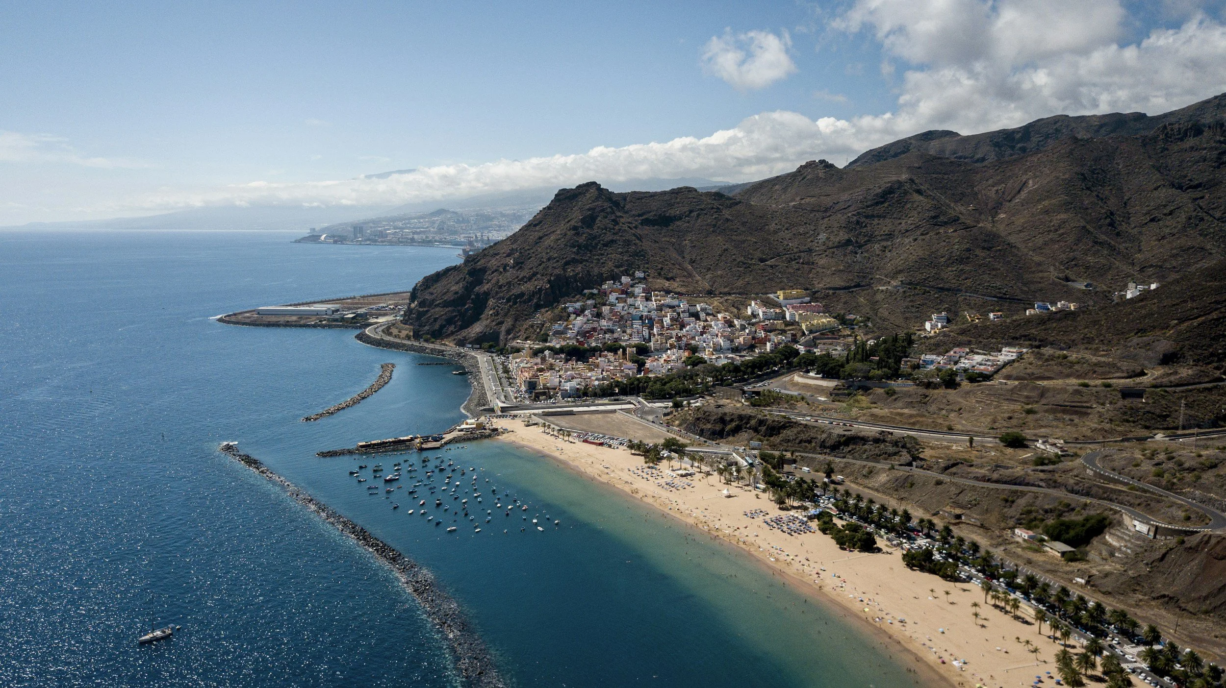 Aerial view of a beach and coastal town with mountains in the background, featuring blue ocean water, sandy beaches, and piers.