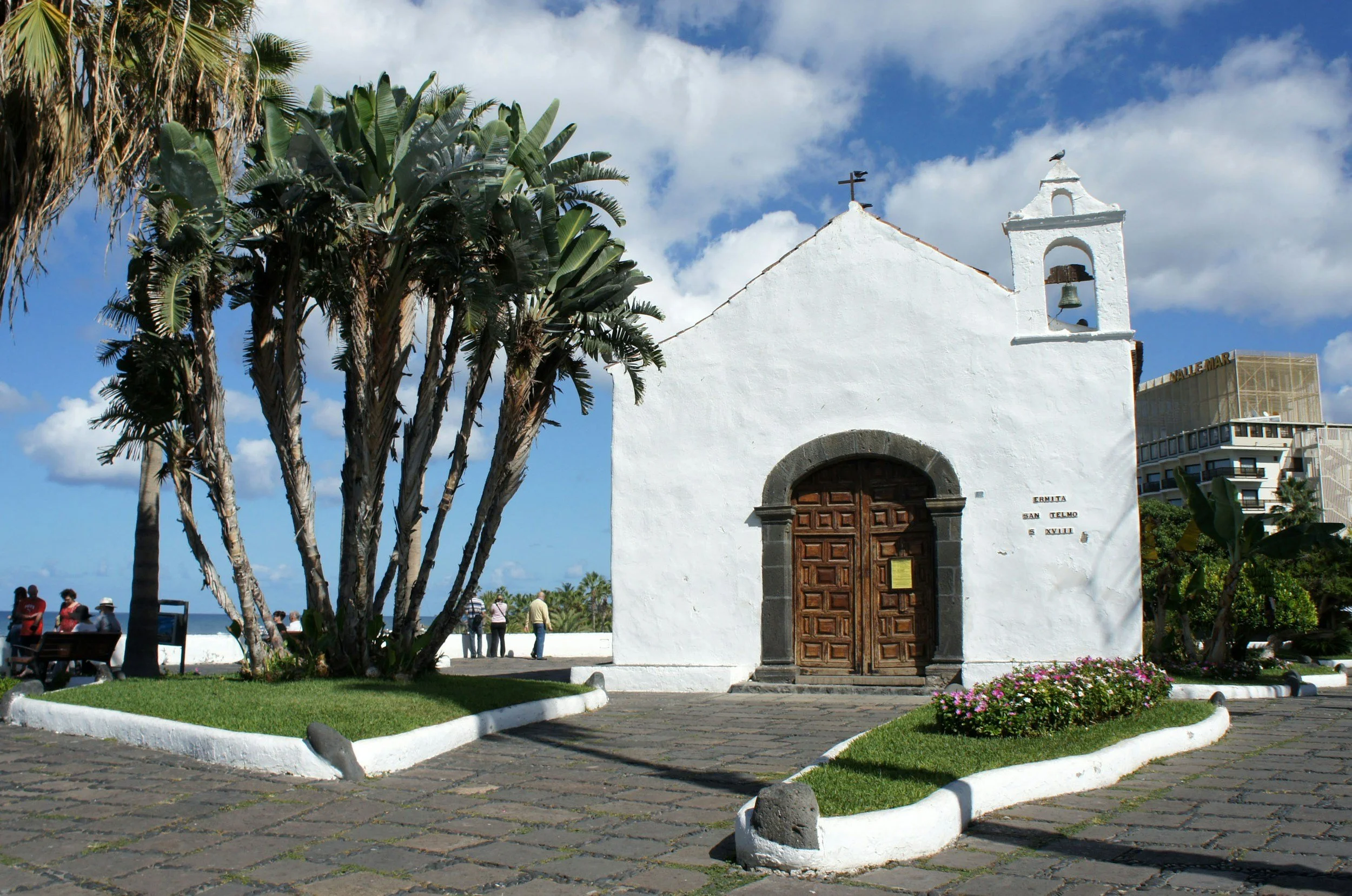 White chapel with wooden door and bell tower, surrounded by palm trees and blue sky.