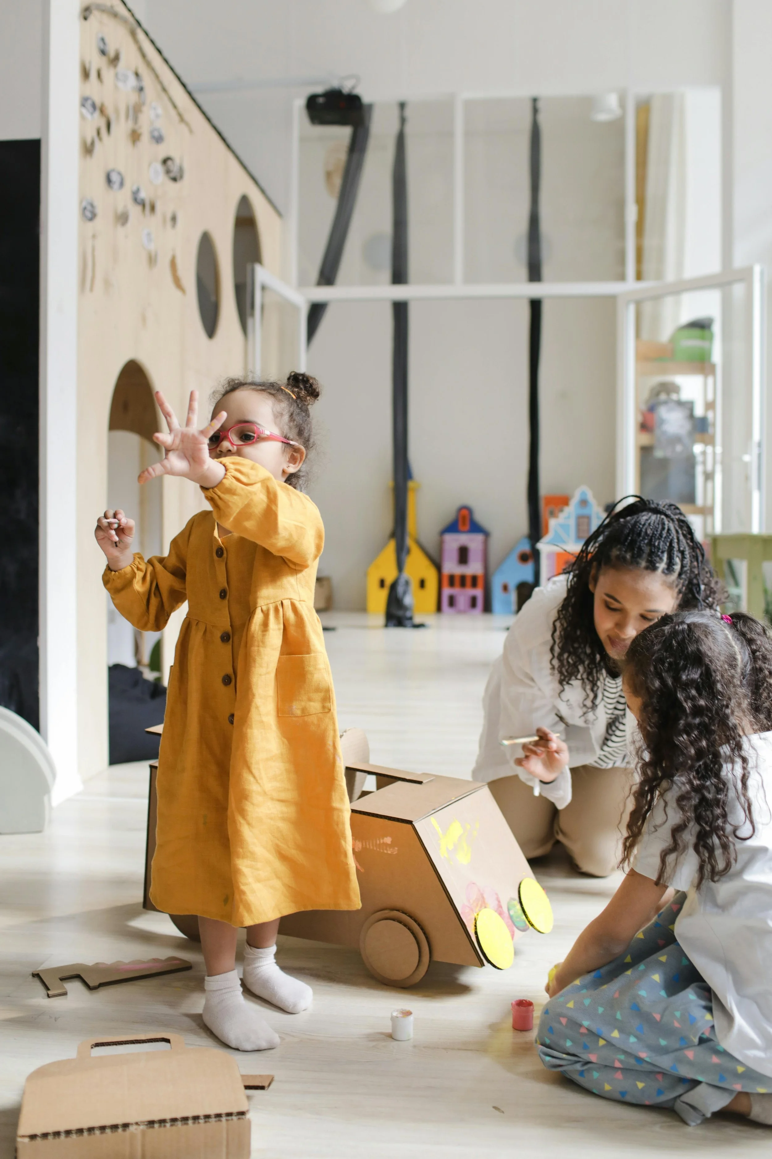 Children playing and crafting with a cardboard cart decorated with colorful stickers, while a woman assists and a girl in an orange dress and pink glasses looks on indoors.