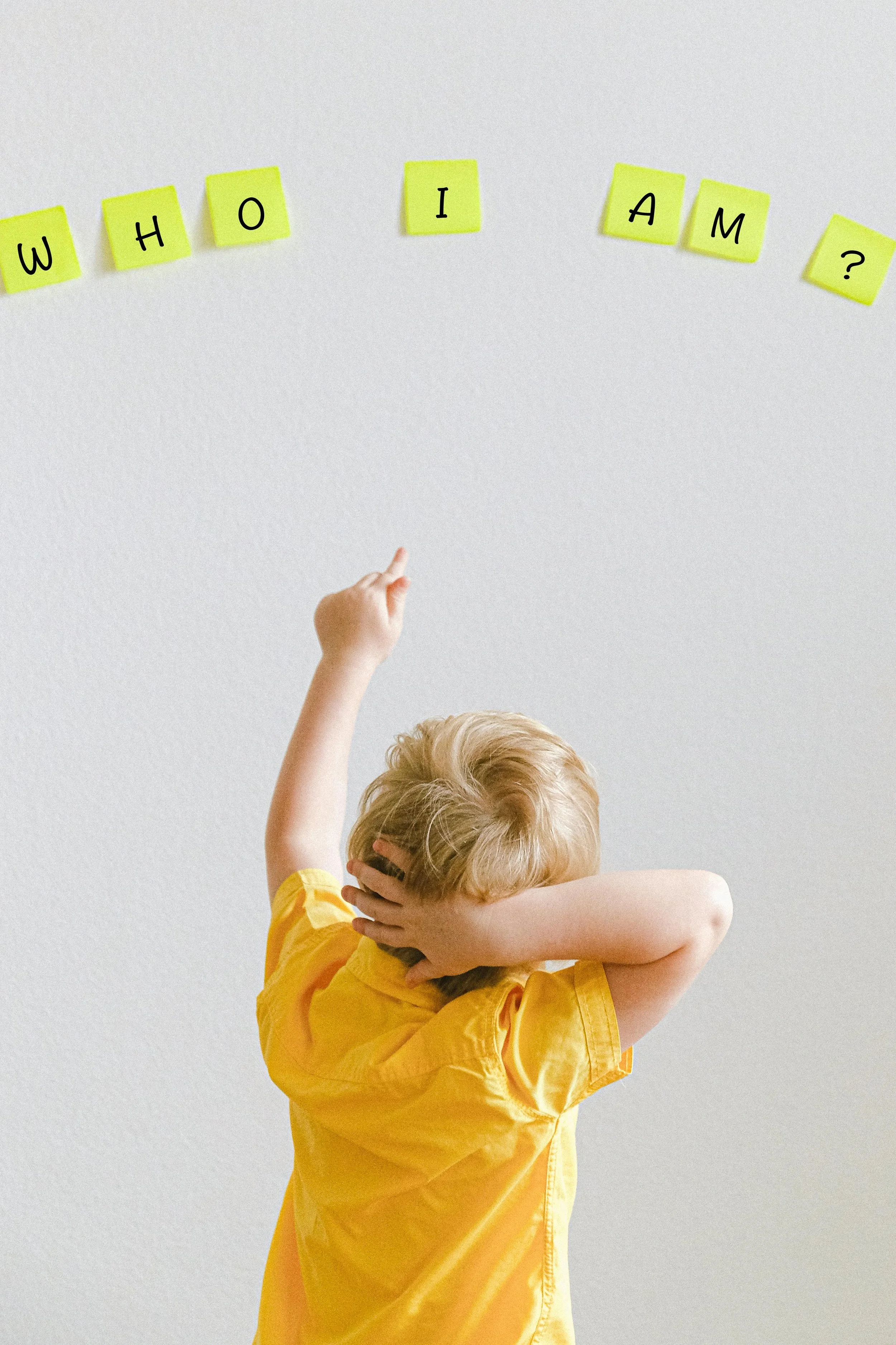 Child in yellow shirt with blonde hair covering ears, standing against a white wall with yellow sticky notes spelling out 'WHO I AM ?'