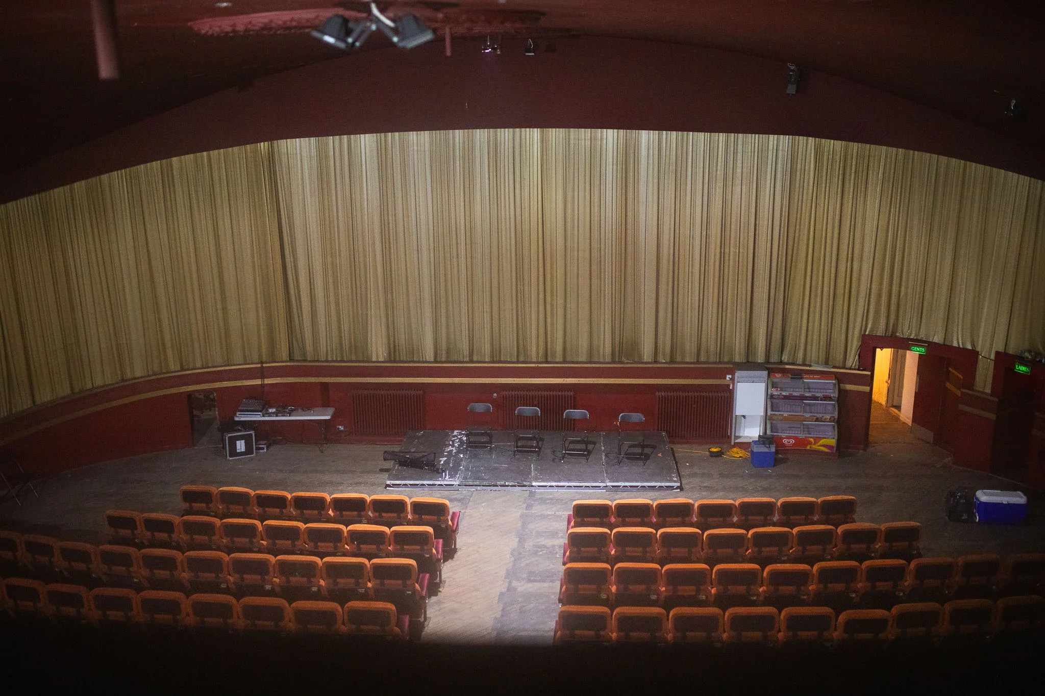 An empty theater stage with chairs and equipment, framed by rows of orange seats, and a large closed yellow curtain behind the stage.