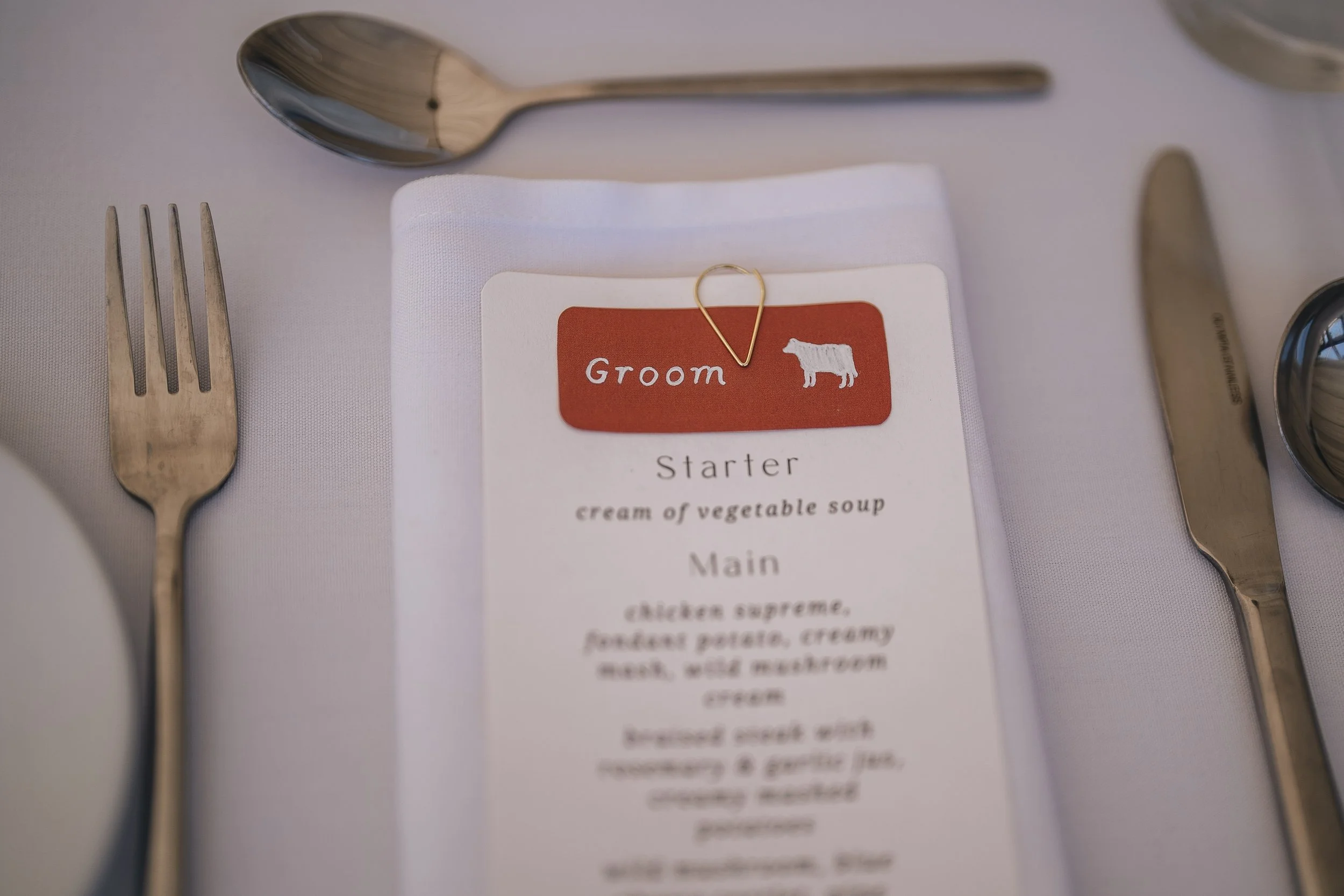 Wedding menu card placed on a white tablecloth with silver fork on the left and silver knife on the right. The menu has a red label with a cow silhouette and the word 'Groom'.