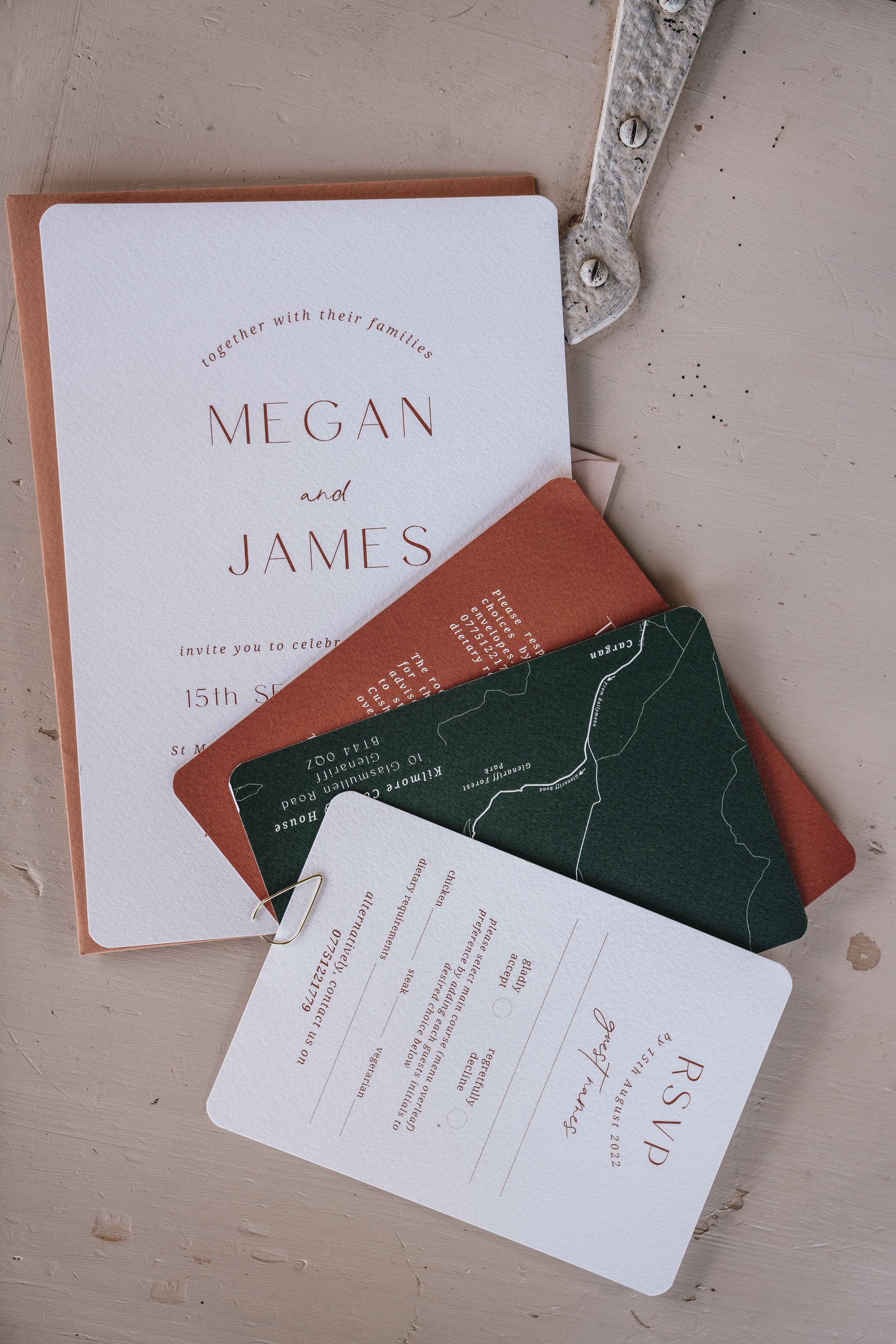 Wedding invitation suite with name Megan and James, RSVP card, map, and additional info on a wooden surface.