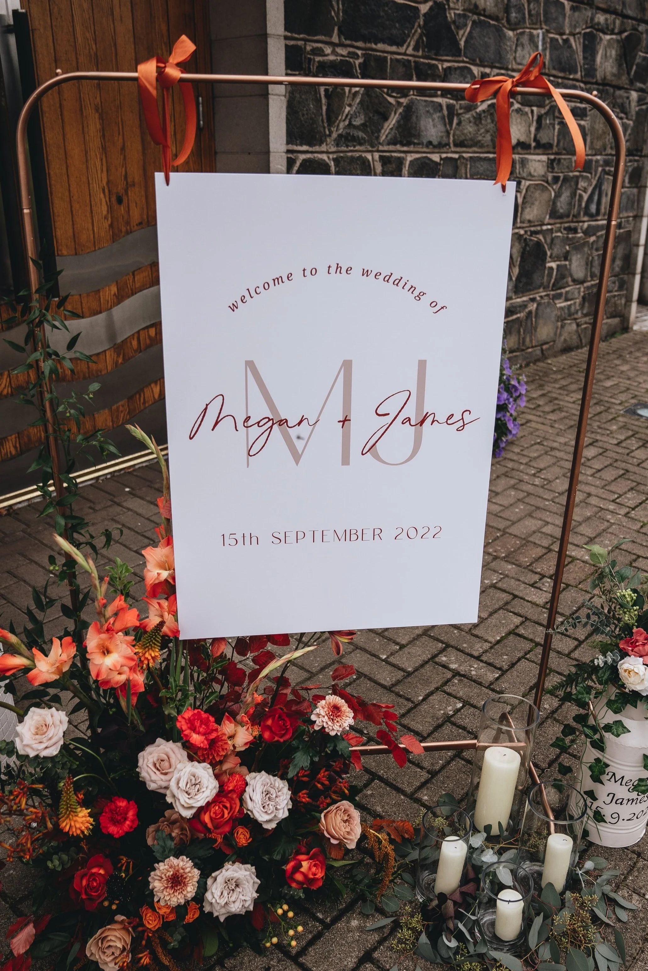 Wedding welcome sign with red, orange and white flowers and candles on a brick sidewalk. The sign reads, "Welcome to the wedding of Megan + James, 15th September 2022."