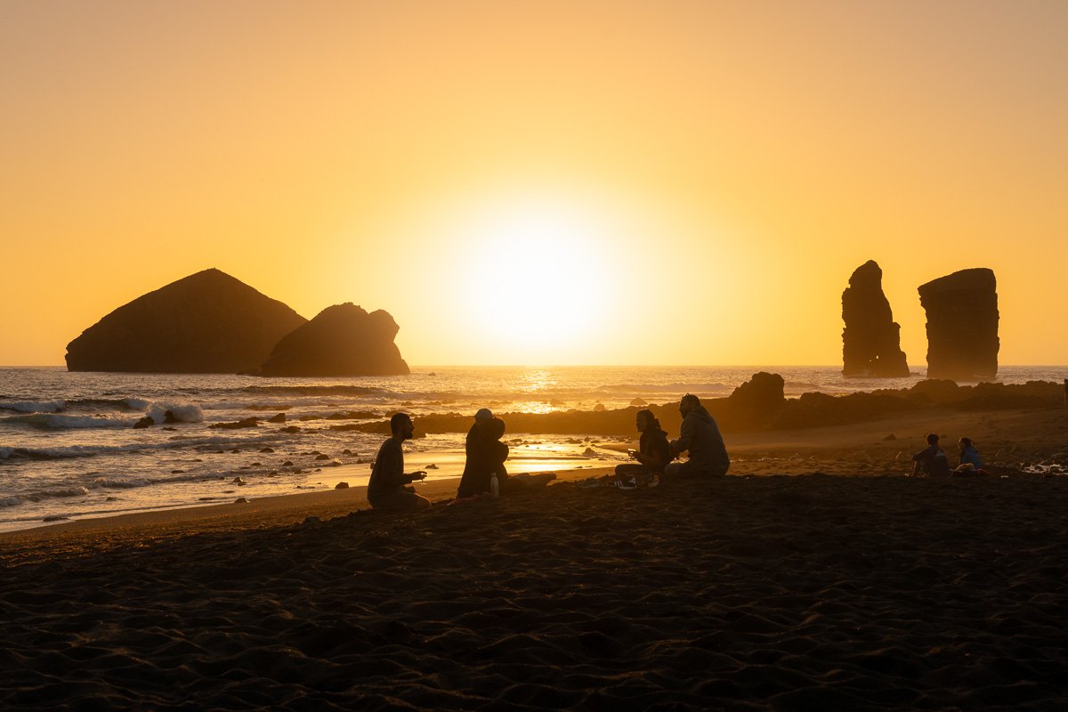 People sitting on a beach at sunset with rock formations in the background.