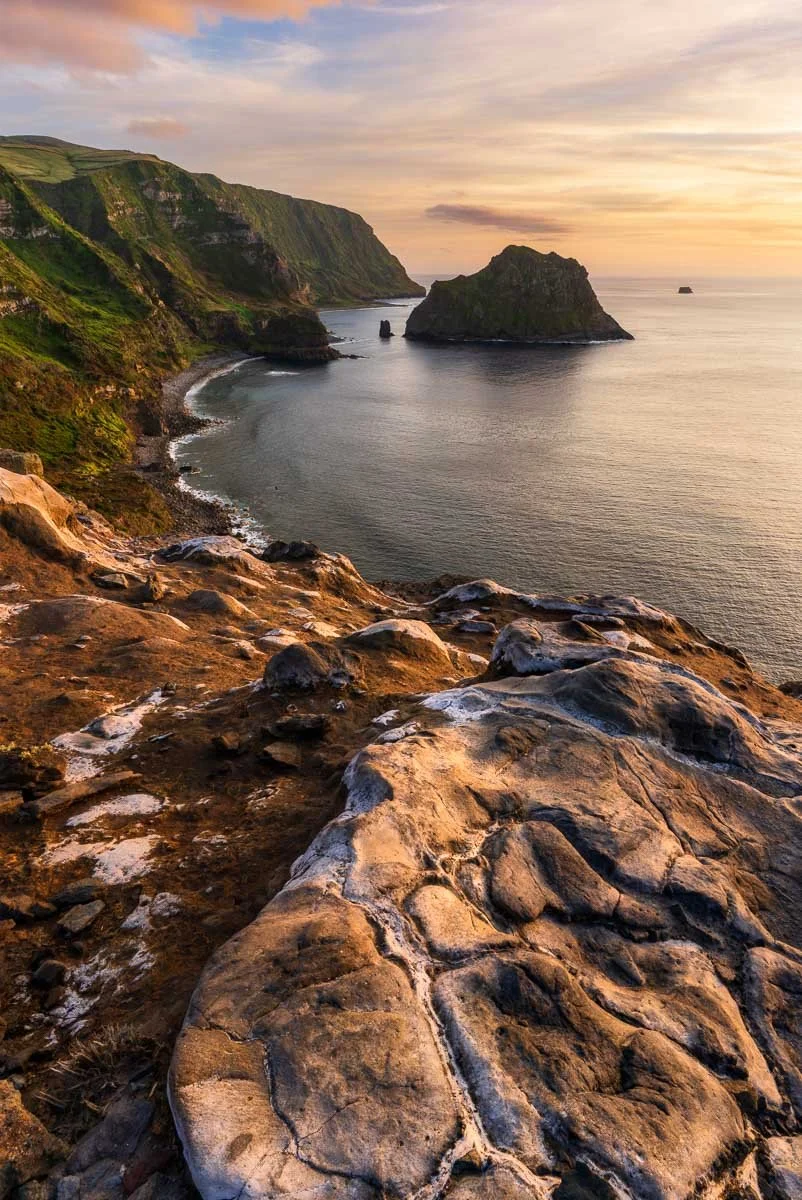 Rocky cliff overlooking a calm ocean with large rocks and green hillside cliffs in the distance during sunset.