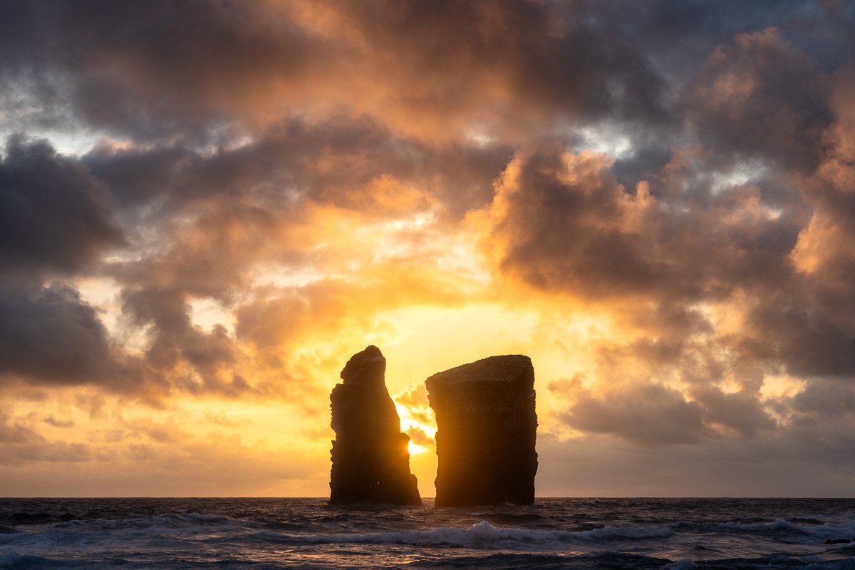 Two large sea stacks rising from the ocean at sunset, with a vibrant orange and yellow sky and scattered dark clouds.