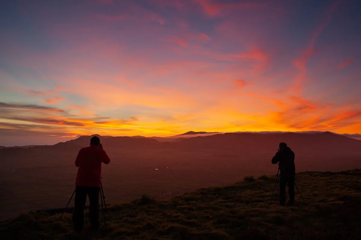 Two photographers standing on a hill during sunset, capturing the colorful sky with mountains in the background.