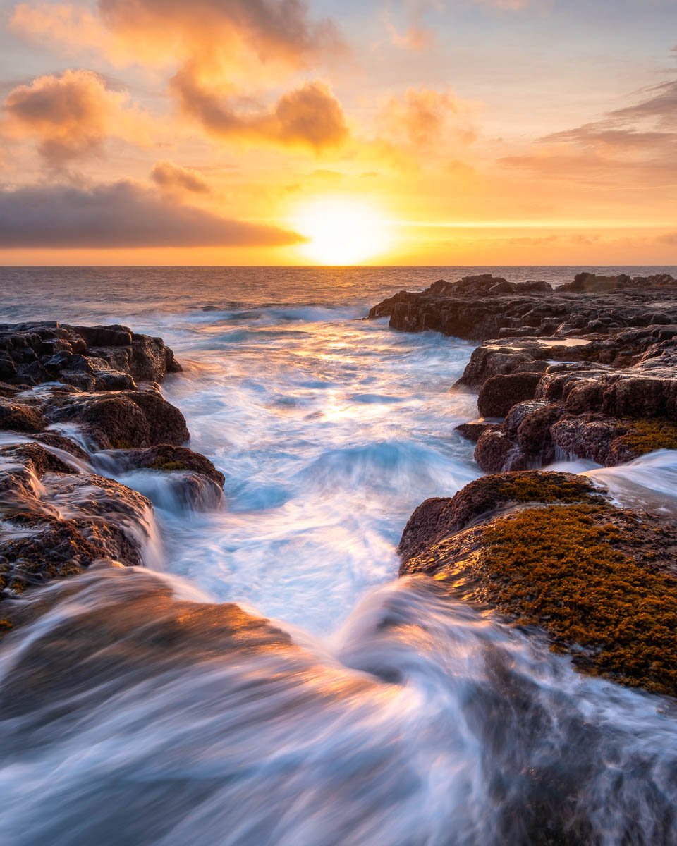 Sunset over the ocean with waves crashing against rocky shoreline