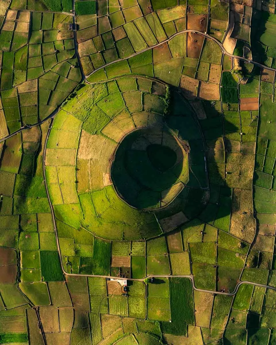 Aerial view of a spiral-shaped green field surrounded by smaller rectangular fields, with a road passing through and a shadow cast over part of the landscape.