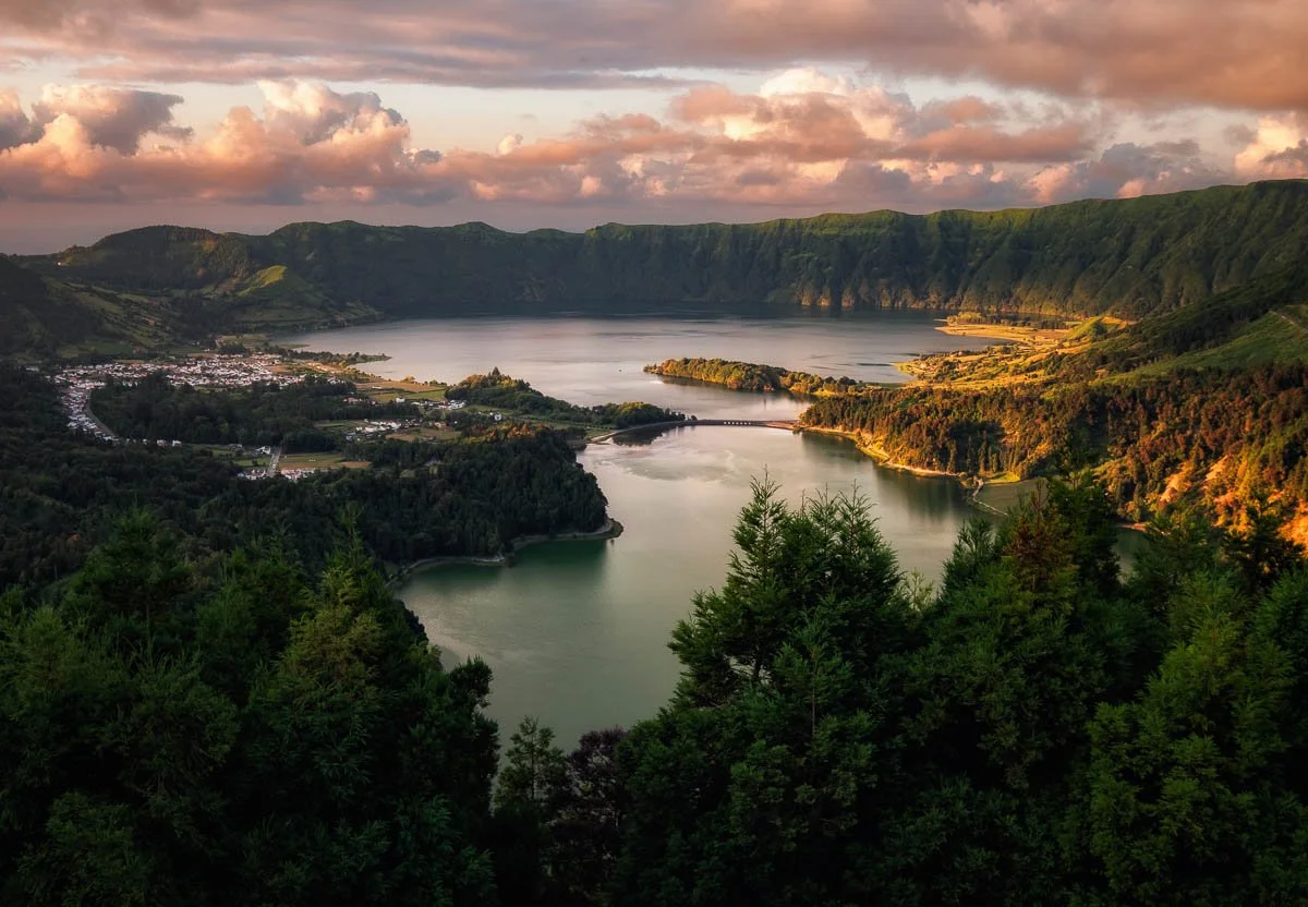 Panoramic view of a lake surrounded by lush green hills and a small town in the distance during sunset.