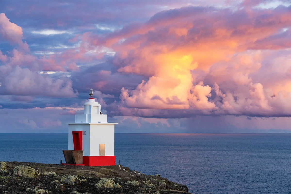 A small white lighthouse with a red door on a rocky coastline, overlooking the ocean at sunset with colorful clouds in the sky.