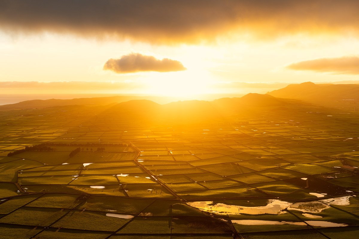 Aerial view of fields at sunset with golden light, scattered water ponds, and distant mountains.