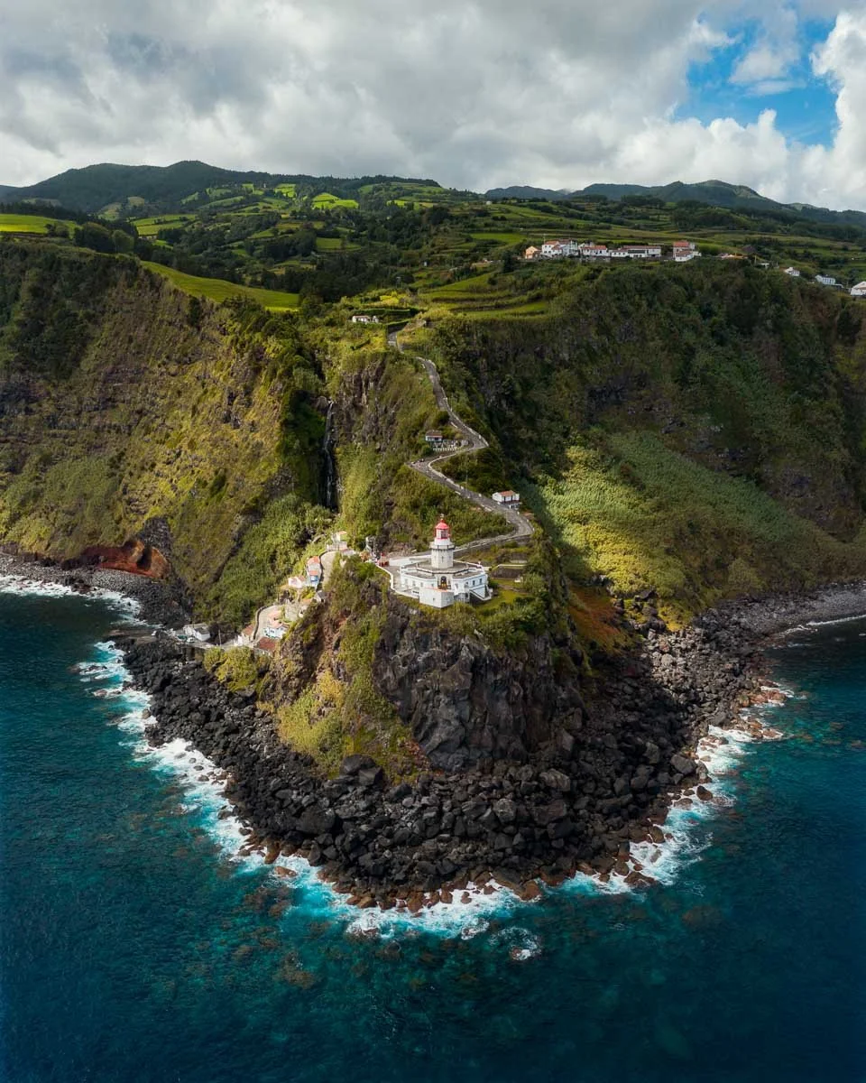 A coastal landscape featuring a white lighthouse on a rocky promontory, with a winding road leading up through green hills and a cliffside, under a partly cloudy sky.