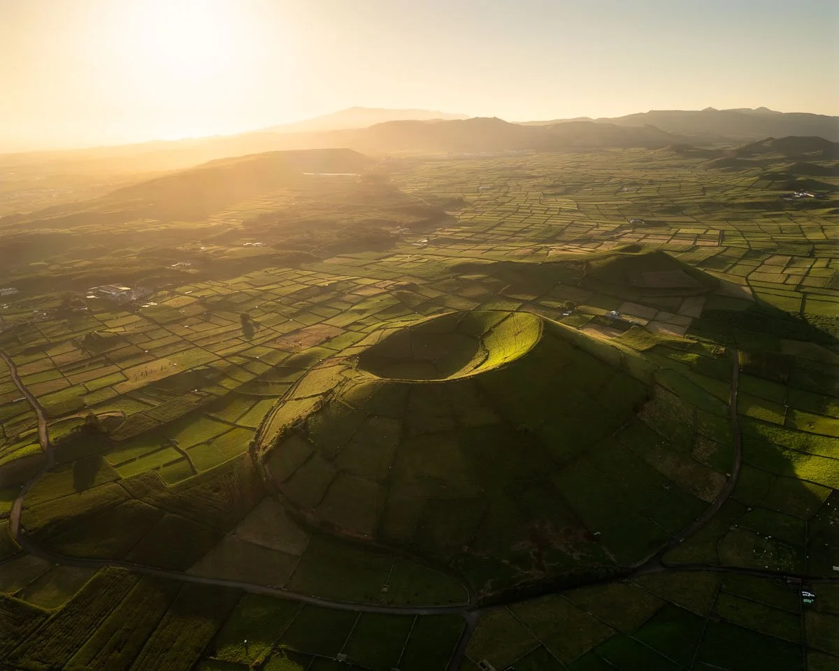 Aerial view of a landscape with a prominent volcanic crater in the center, surrounded by patchwork farmland, with mountains in the background during sunset.