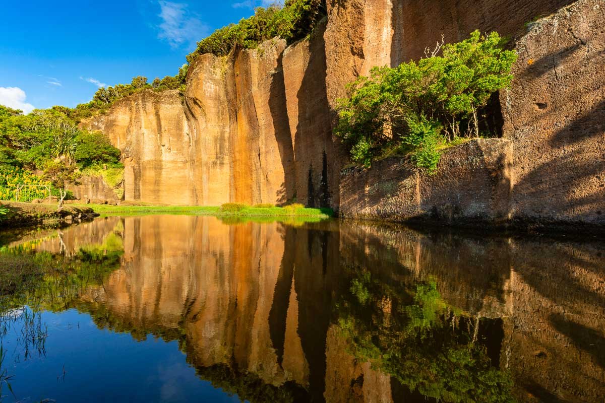 Cliffs beside a river with green trees and grass, clear blue sky, and their reflections in the water.