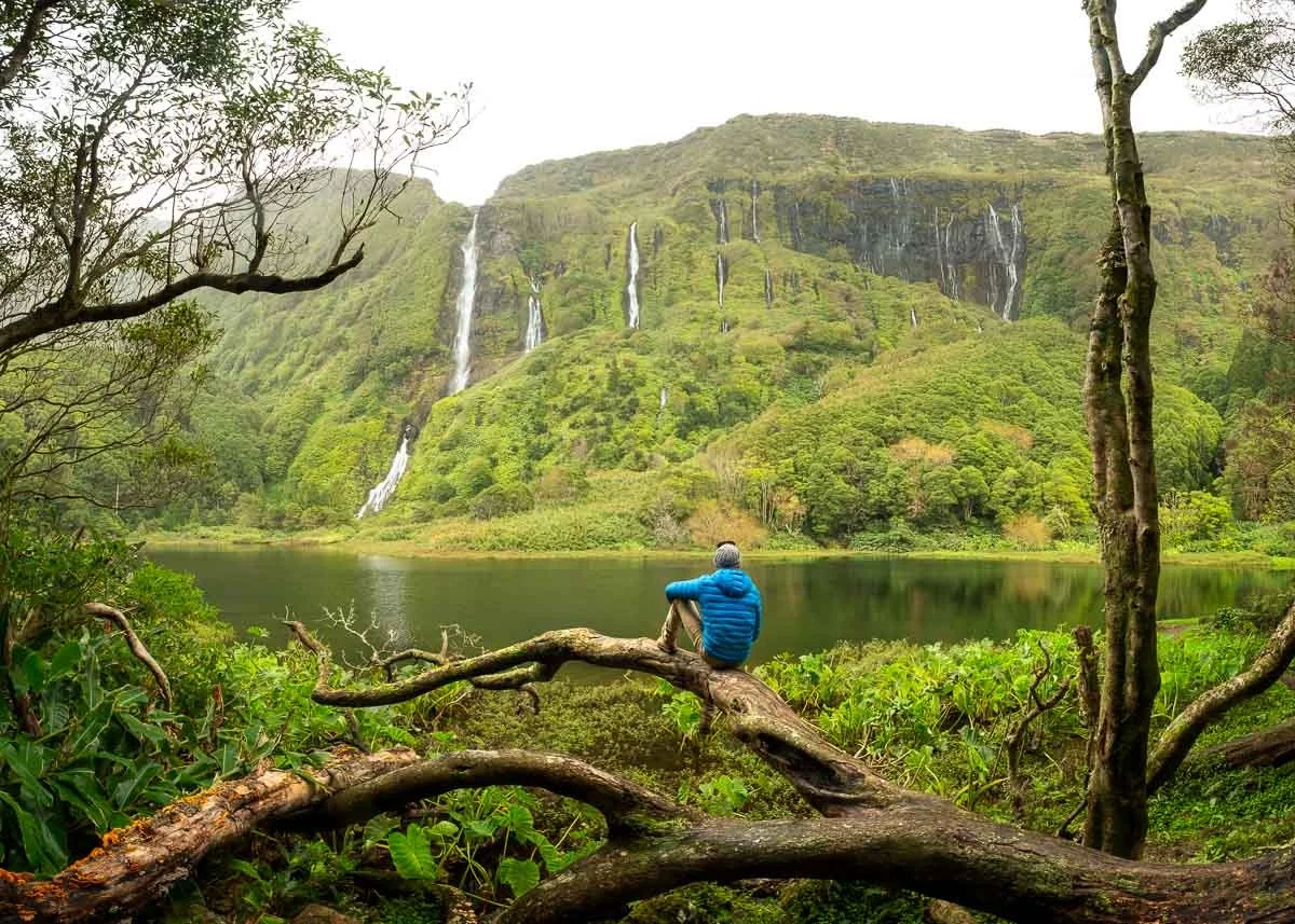 A person with a blue jacket and gray hat sits on a fallen tree overlooking a lake with a lush green mountain and multiple waterfalls in the background.