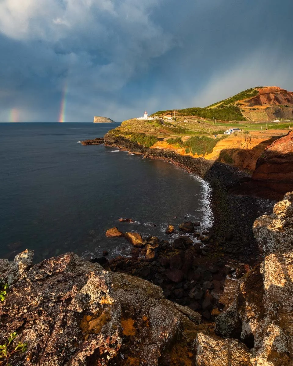 Cliffs overlooking the ocean with a lighthouse on top, stormy sky, rainbow in the distance, rocky shoreline, and a small beach.
