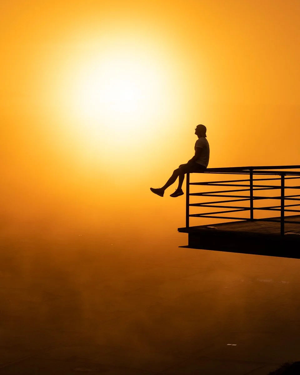 A person sitting on the edge of a pier or balcony with their legs hanging, silhouetted against a bright, setting sun in an orange-yellow sky.