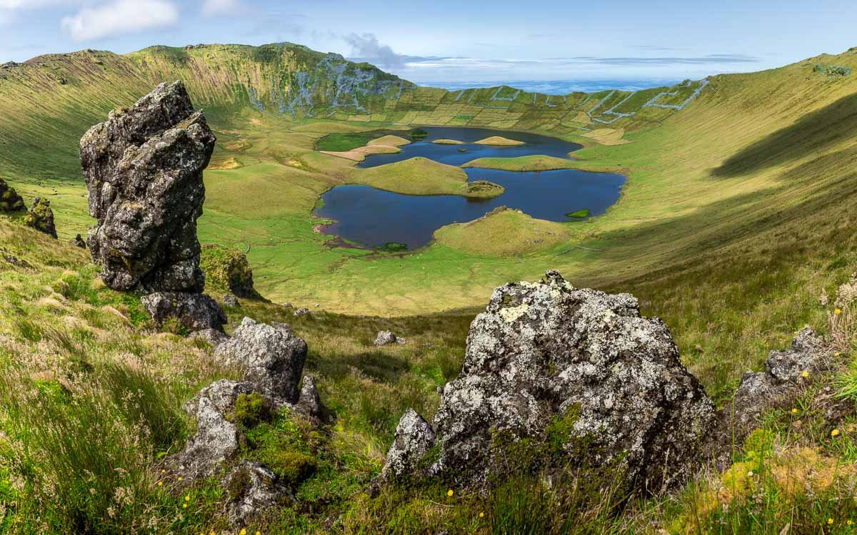 A volcanic crater filled with blue lakes and green grass, surrounded by rocky formations and hills, with a partly cloudy sky in the background.