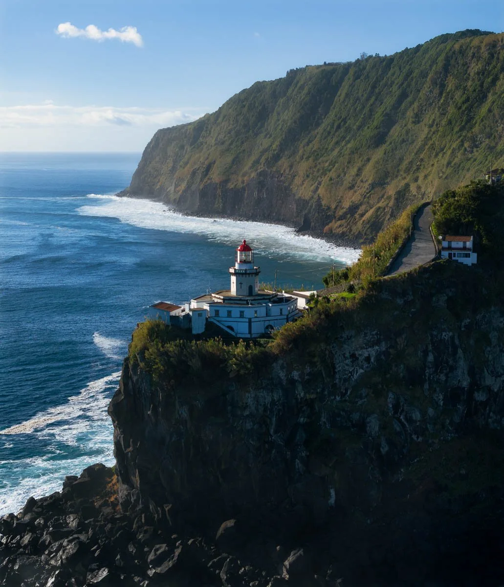 Cliffside lighthouse on a rocky coast, overlooking the ocean with waves and a hillside covered in greenery.