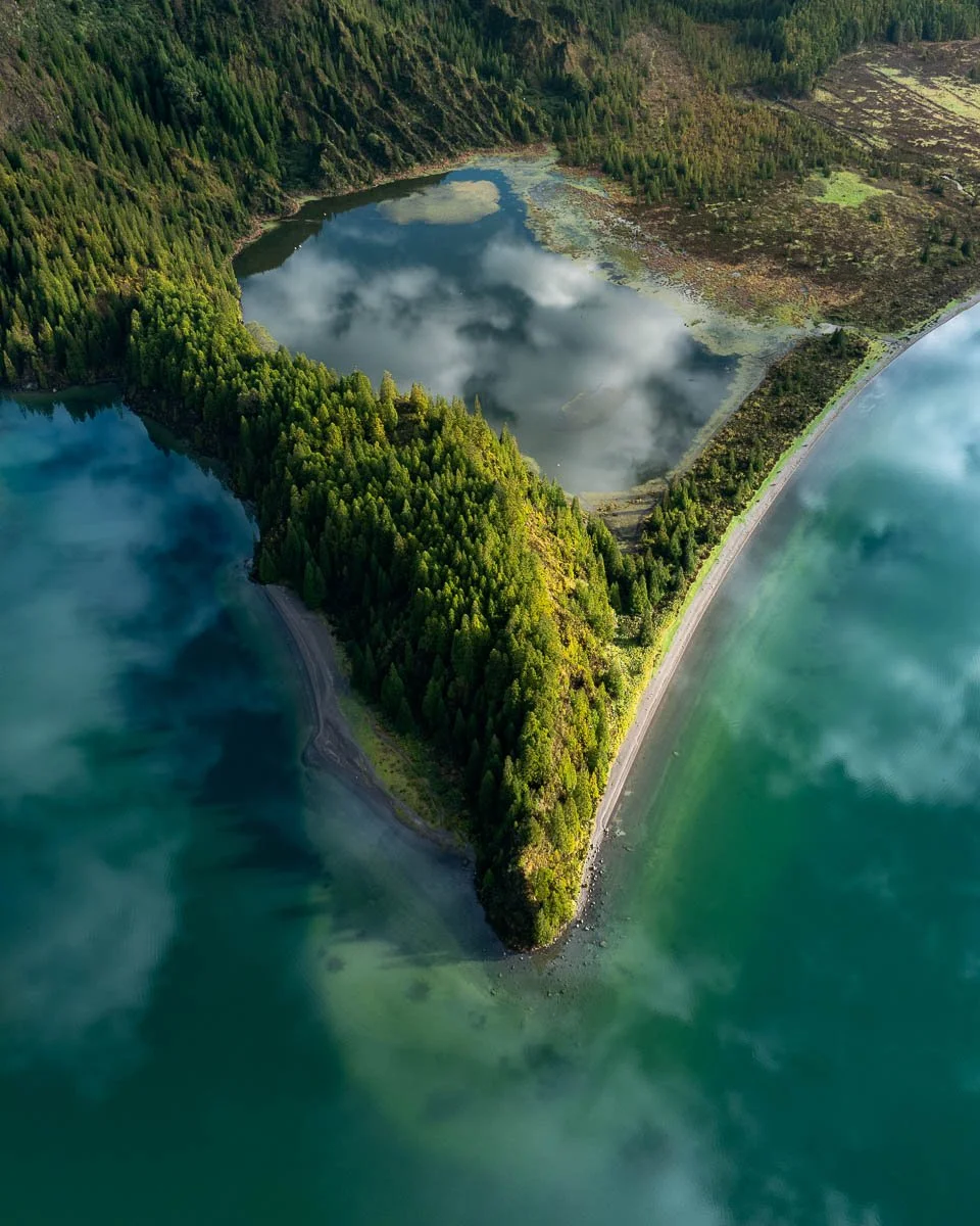 Aerial view of a small, forested peninsula surrounded by water with reflections of clouds, and a lake with a mix of trees and open land in the background.