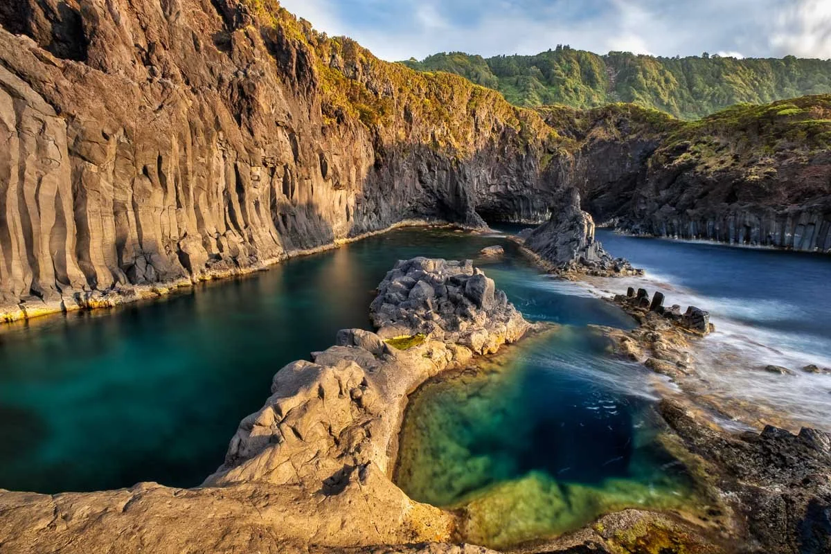 Coastal landscape with rocky shoreline, tide pools, and cliffs covered in greenery under a blue sky.