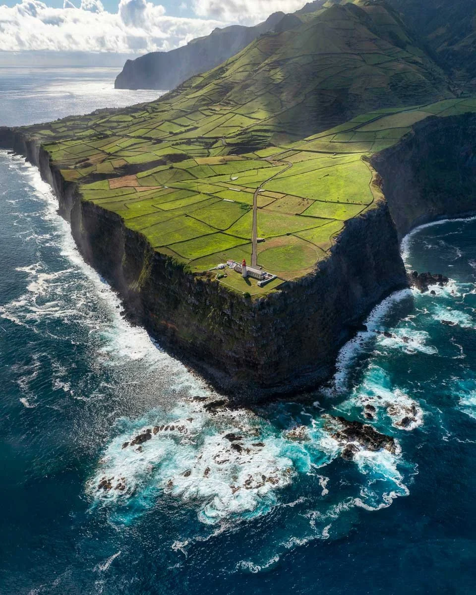 Aerial view of a green, terraced landscape on a coastal island with cliffs and waves crashing below.