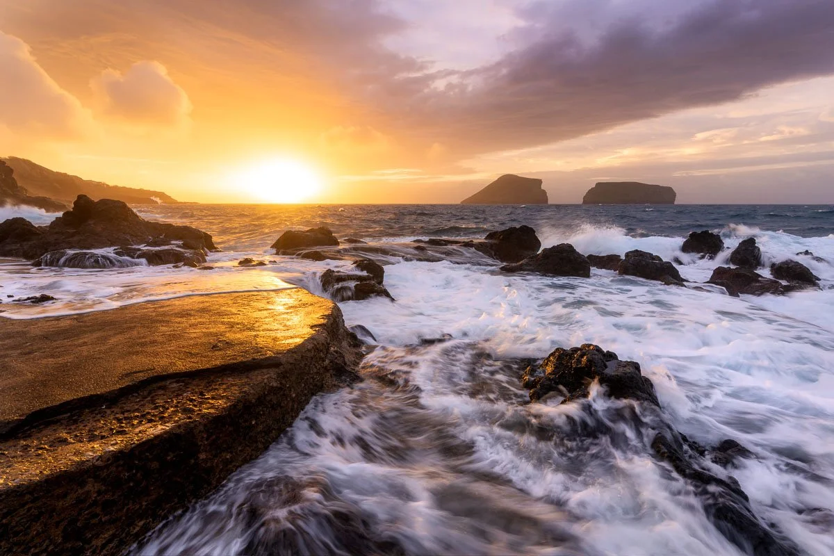 A vibrant sunset over the ocean with rocky shoreline and two distant islands on the horizon.