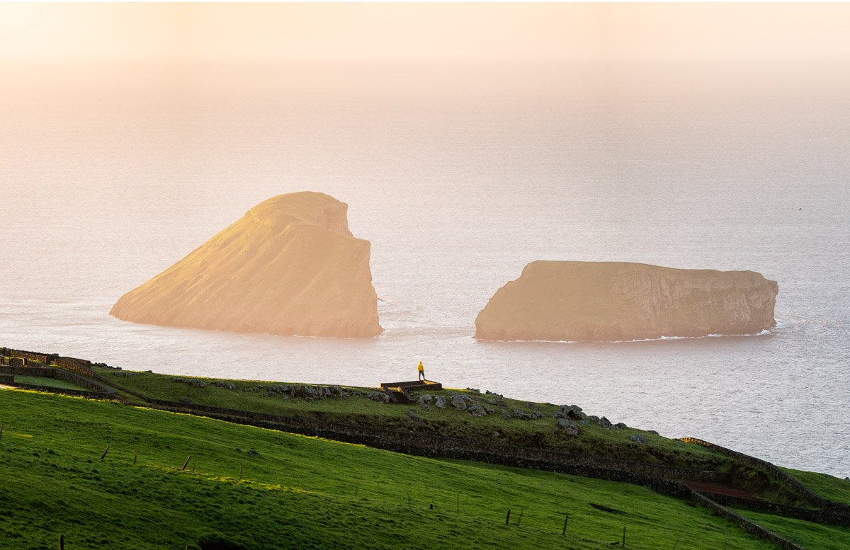 A person standing on a grassy hillside overlooking the ocean with two large rocky islands in the distance, during sunset.