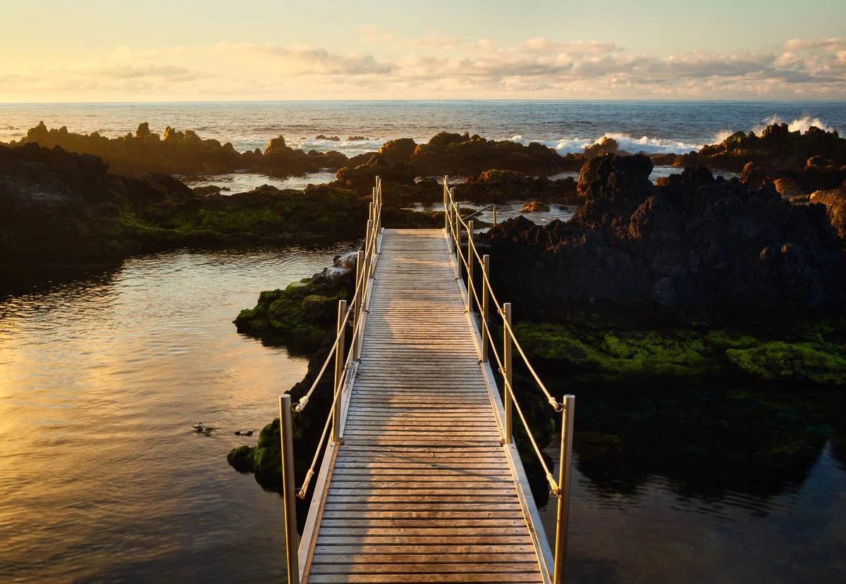 A wooden dock extends over rocky ocean tide pools at sunset, with waves crashing in the background and the sky partly cloudy.