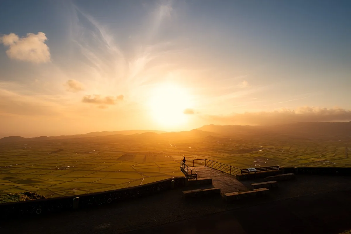 A person standing on a viewing platform overlooking a vast, flat landscape during sunset with a partly cloudy sky.