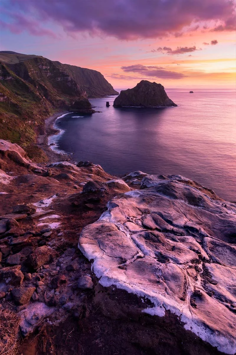 Sunset over the ocean with large rocks and cliffs along the coastline, displaying pink, purple, and orange hues in the sky and reflected on the water.