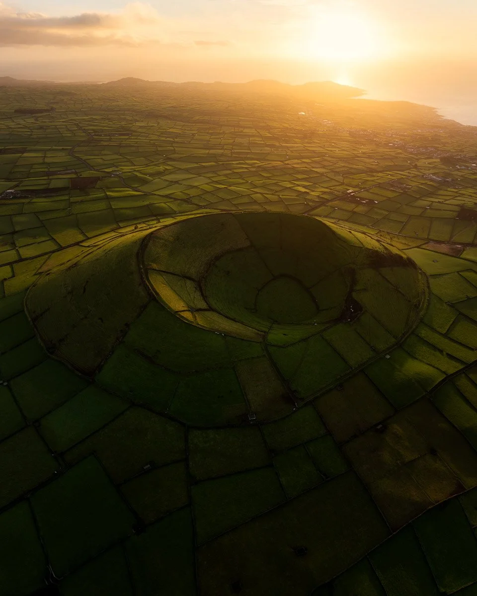 An aerial view of a large, circular, terraced field carved into the landscape at sunset, with patchwork green fields and a rural area in the background.