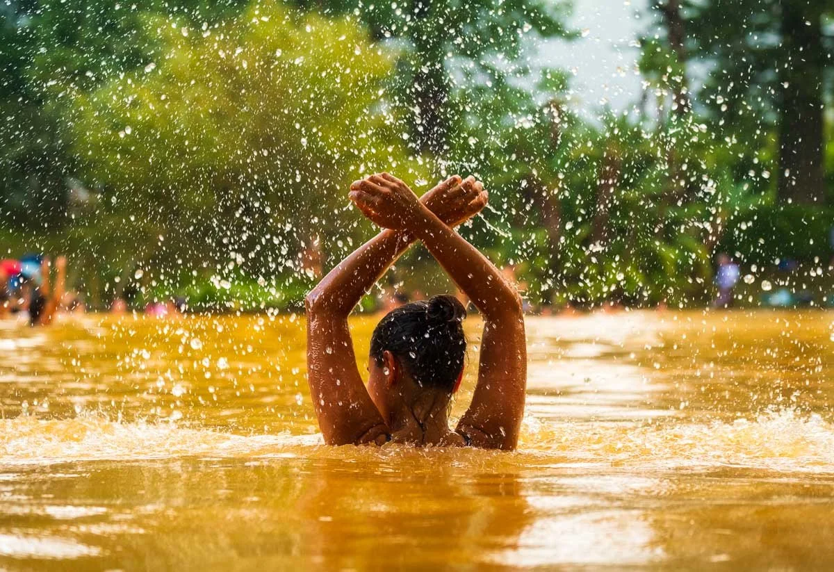 A person swimming in muddy water, with arms crossed above their head, water splashing around, and a background with green trees.
