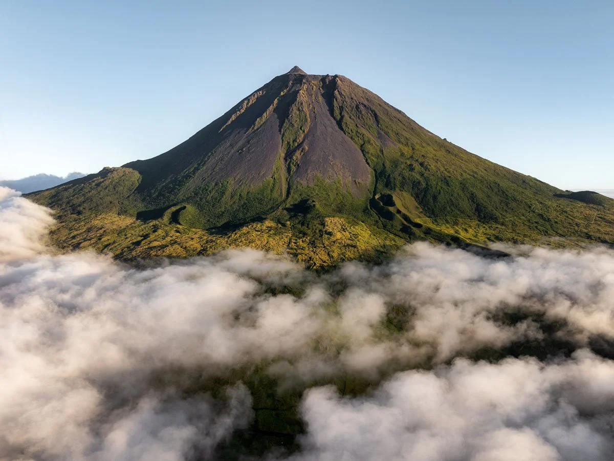 A tall volcanic mountain with a green and dark rocky surface, surrounded by clouds at its base, under a clear blue sky.